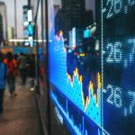 Een scherm met daarop een index van een aandelenbeurs, met mensen op de achtergrond.