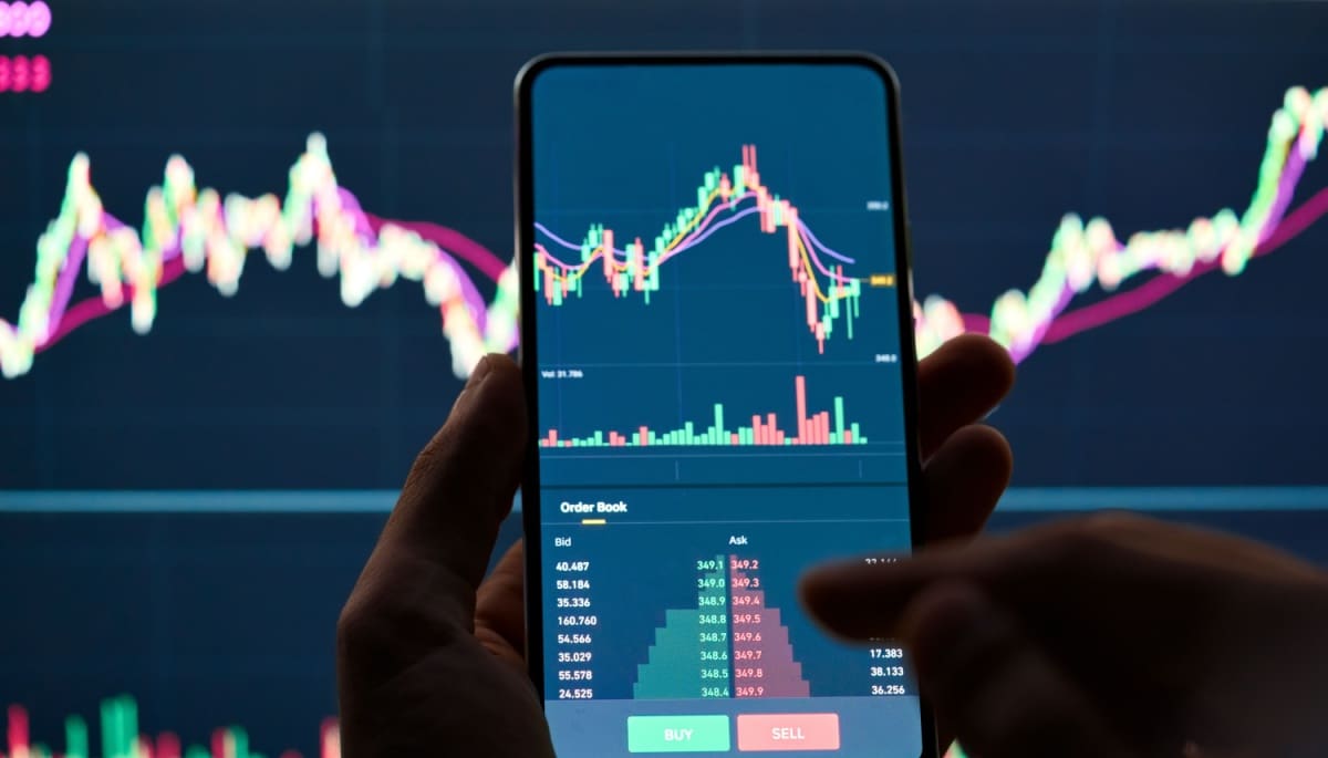 Een crypto handelaar analyseert de bitcoin koers op zijn mobiel, met op de achtergrond een groter scherm met marktdata