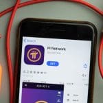 Een afbeelding van de Pi network app op een mobiele telefoon, met daaromheen kantoorspullen. Pi Network (PI) maakt minen op de telefoon mogelijk, en heeft 60 miljoen gebruikers