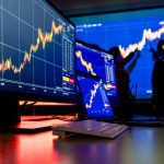 Een afbeelding van meerdere beeldschermen met koersen van crypto en aandelen en op de achtergrond beleggers die een koers analyseren