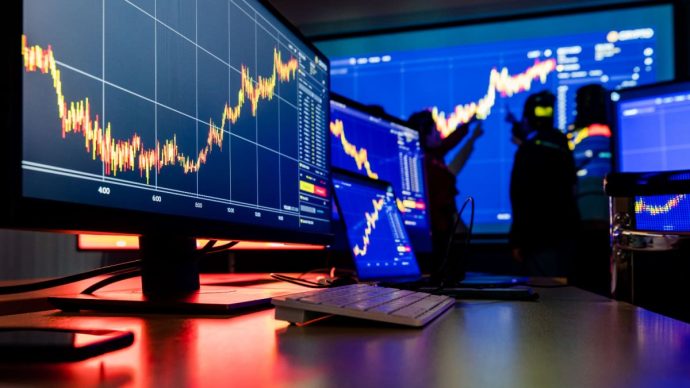 Een afbeelding van meerdere beeldschermen met koersen van crypto en aandelen en op de achtergrond beleggers die een koers analyseren