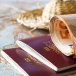 Een afbeelding met 50 euro bankbiljetten op een twee paspoorten met een kaart en een vakantiehoed op de achtergrond