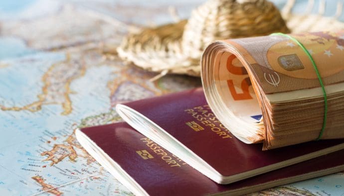 Een afbeelding met 50 euro bankbiljetten op een twee paspoorten met een kaart en een vakantiehoed op de achtergrond