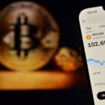Een afbeelding van een mobiele telefoon met daarop de bitcoin prijs boven de $100.000, met op de achtergrond een BTC munt