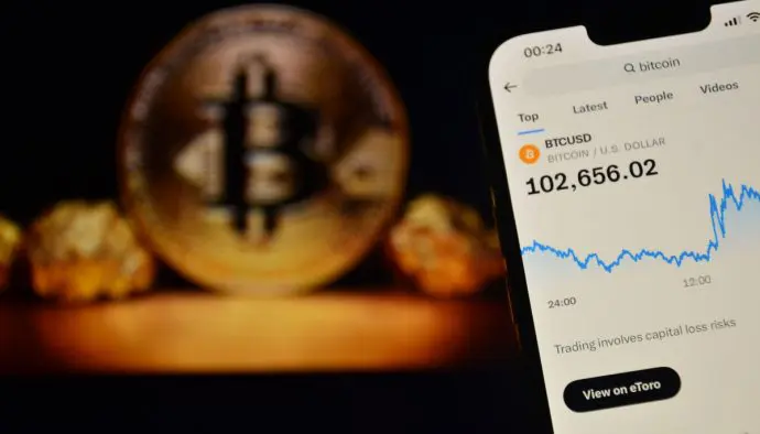 Een afbeelding van een mobiele telefoon met daarop de bitcoin prijs boven de $100.000, met op de achtergrond een BTC munt