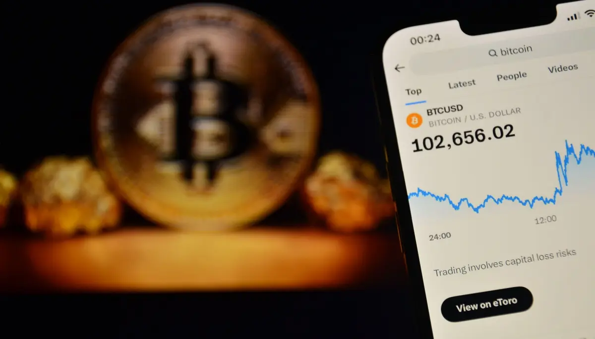 Een afbeelding van een mobiele telefoon met daarop de bitcoin prijs boven de $100.000, met op de achtergrond een BTC munt