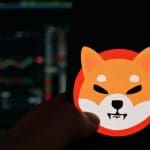 Het logo van de memecoin shiba inu (SHIB) op een mobiele telefoon met op de achtergrond een koers in candle chart format, op een cryptobeurs