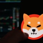 Het logo van de memecoin shiba inu (SHIB) op een mobiele telefoon met op de achtergrond een koers in candle chart format, op een cryptobeurs