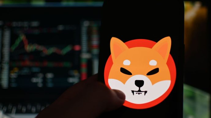 Het logo van de memecoin shiba inu (SHIB) op een mobiele telefoon met op de achtergrond een koers in candle chart format, op een cryptobeurs