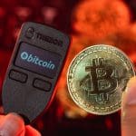 Een afbeelding van een Trezor crypto hard wallet met ernaast een bitcoin (BTC) munt en op de achtergrond een rode kleur