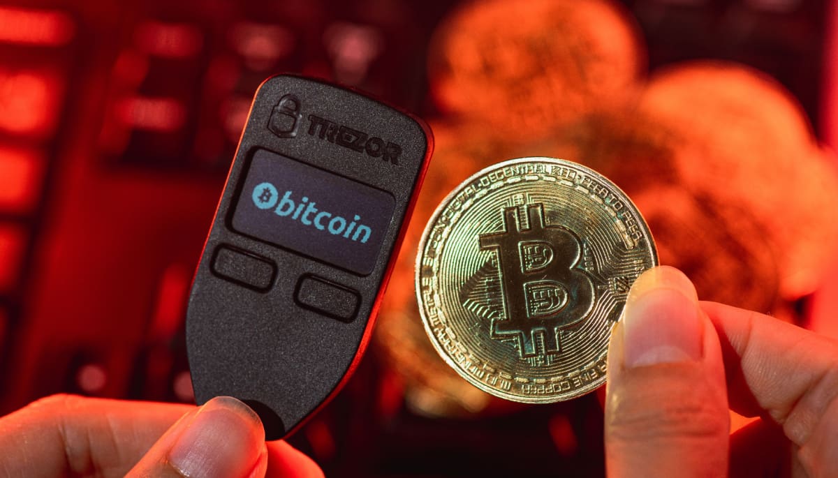 Een afbeelding van een Trezor crypto hard wallet met ernaast een bitcoin (BTC) munt en op de achtergrond een rode kleur