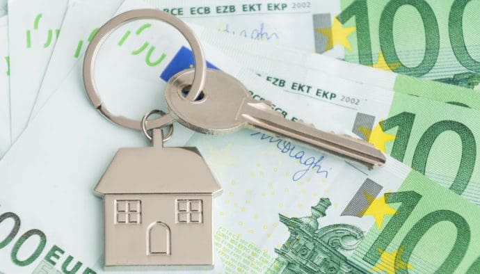 Een afbeelding van een huissleutel met een huis eraan, en op de achtergrond euro bankbiljetten, wat de woningmarkt en de bijbehorende stijgende prijzen moet representeren