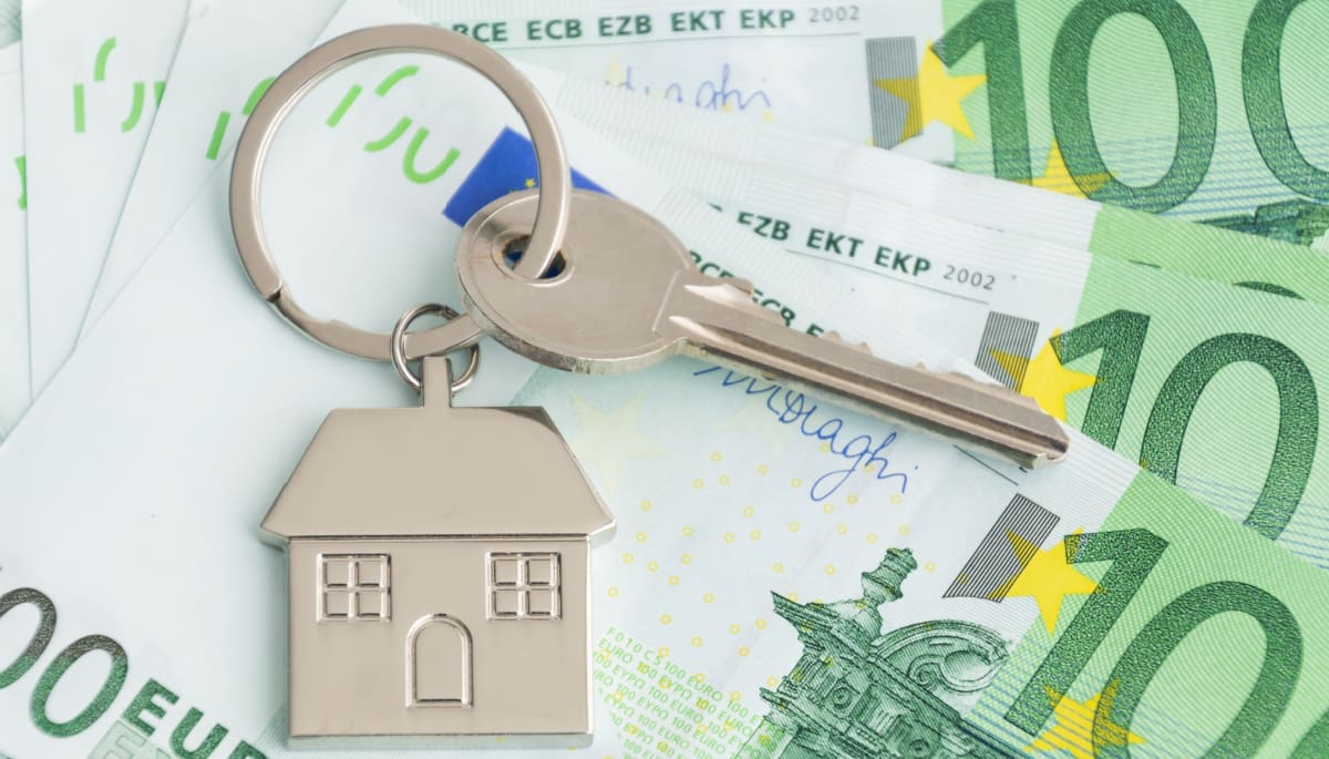 Een afbeelding van een huissleutel met een huis eraan, en op de achtergrond euro bankbiljetten, wat de woningmarkt en de bijbehorende stijgende prijzen moet representeren