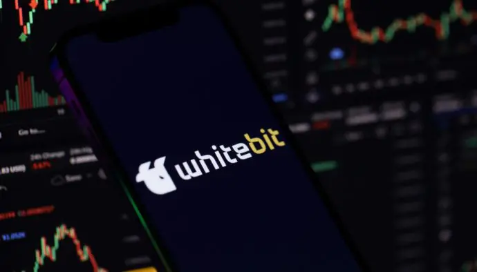 Een afbeelding van whitebit, een cryptomunt afkomstig van een grote crypto exchange. Het logo van whitebit staat op een mobiel, en op de achtergrond staan grafieken