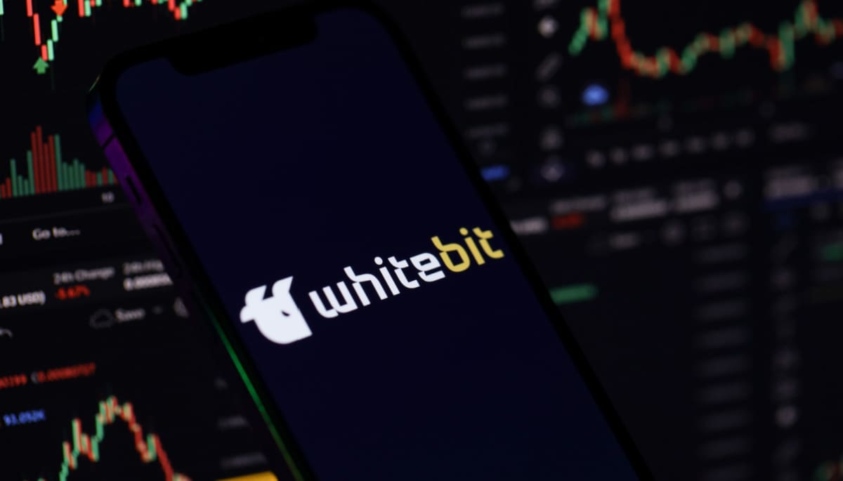 Een afbeelding van whitebit, een cryptomunt afkomstig van een grote crypto exchange. Het logo van whitebit staat op een mobiel, en op de achtergrond staan grafieken