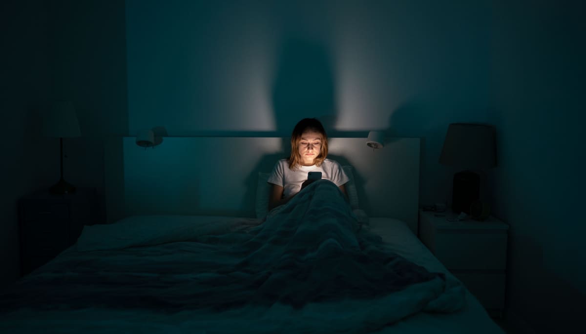 Een afbeelding met een vermoeide vrouw die 's avonds laat of 's nachts op de mobiel zit, omdat slapen moeilijk gaat