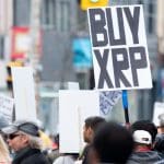 Een afbeelding van een protest waarbij iemand een bord met 'koop XRP', ookwel Ripple genoemd, ophoudt