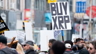 Een afbeelding van een protest waarbij iemand een bord met 'koop XRP', ookwel Ripple genoemd, ophoudt
