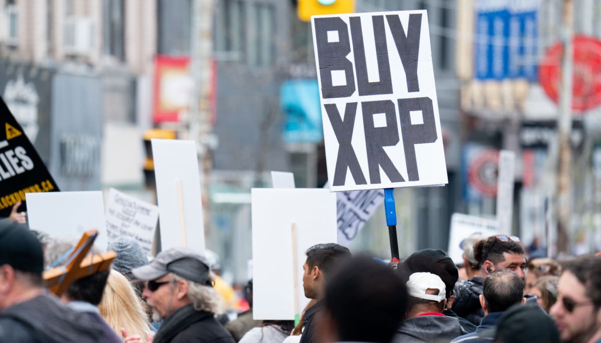 Een afbeelding van een protest waarbij iemand een bord met 'koop XRP', ookwel Ripple genoemd, ophoudt