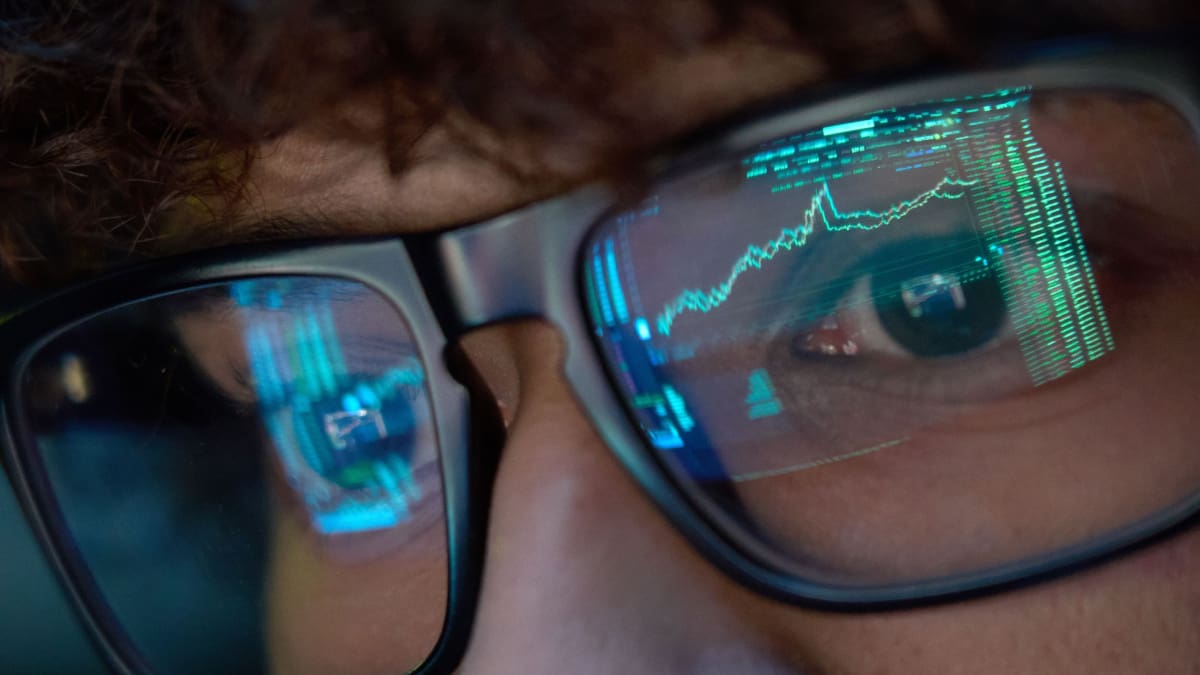 Een close up afbeelding van een crypto handelaar met koersen en grafieken van een cryptobeurs als weerspiegeling op zijn bril