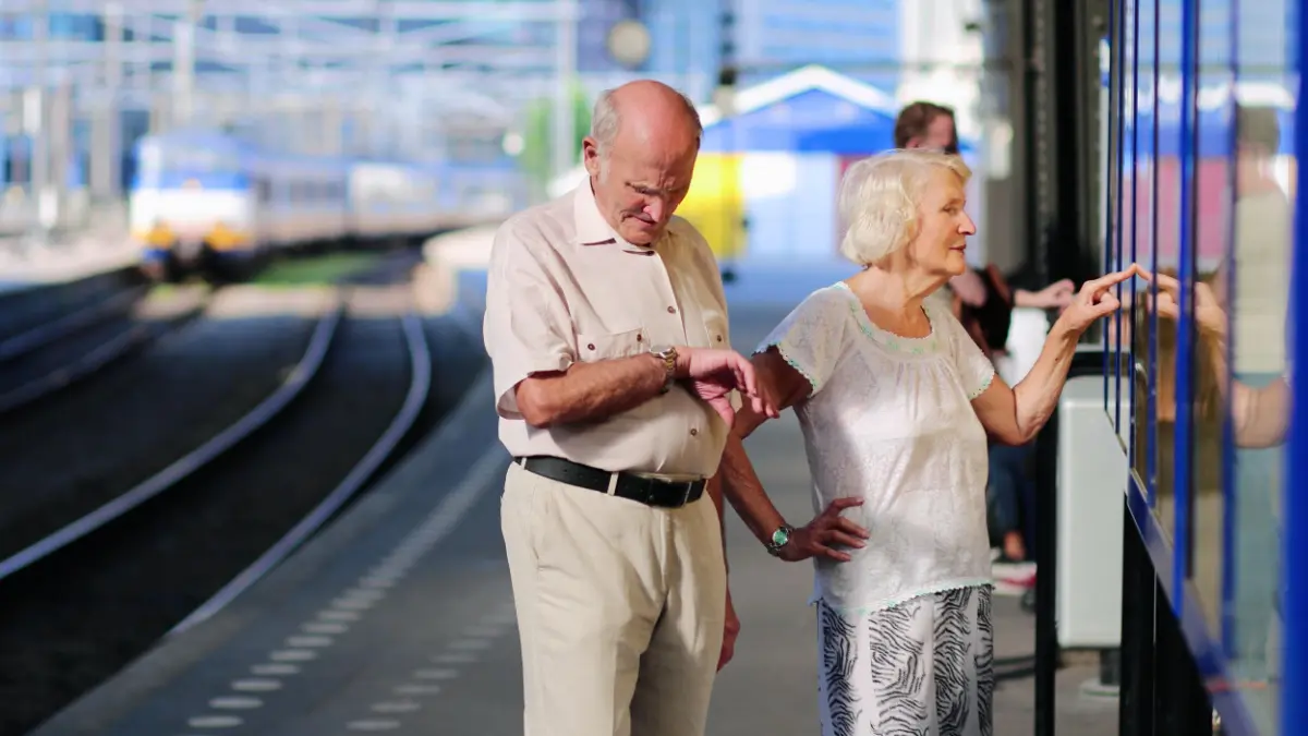 Een afbeelding van een ouder koppel op een treinstation die aan het wachten zijn op het openbaar vervoer