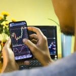 Een afbeelding van een stijgende crypto koers op een mobiele telefoon die door een amateur handelaar wordt vastgehouden
