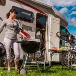 Een afbeelding van een vrouw die aan het barbecueën is op een camping met achter haar een kampeerwagen