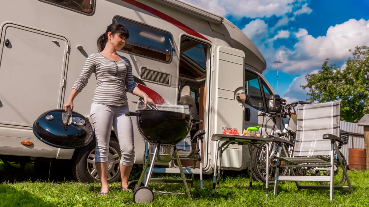 Een afbeelding van een vrouw die aan het barbecueën is op een camping met achter haar een kampeerwagen