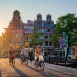 Een afbeelding van mensen die fietsen over een brug in Amsterdam met grachtenhuizen en een zonsondergang op de achtergrond