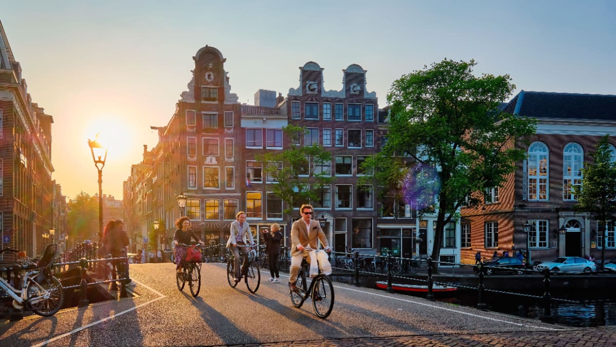 Een afbeelding van mensen die fietsen over een brug in Amsterdam met grachtenhuizen en een zonsondergang op de achtergrond