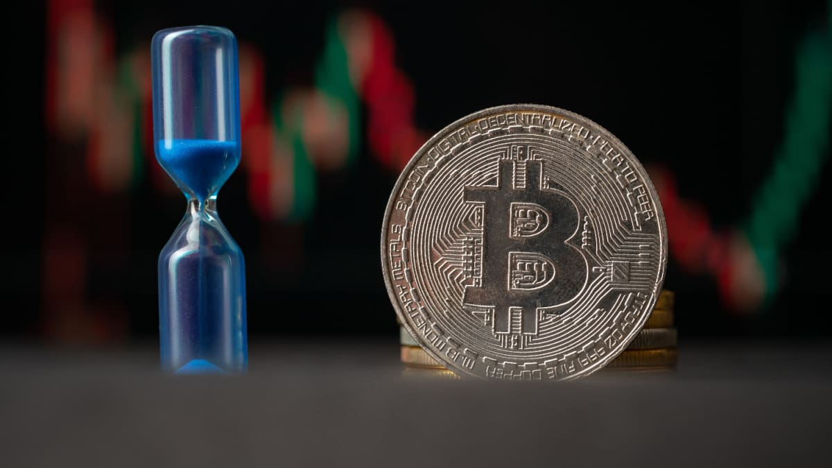 Een afbeelding van een bitcoin (BTC) munt met daarnaast een zandloper en op de achtergrond een grafiek van een volatiele koers