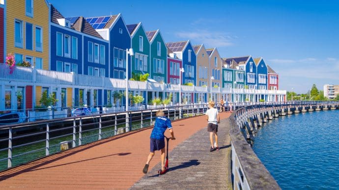 Een afbeelding van twee kinderen die spelen met een step langs het water langs een rij typisch Nederlandse huishoudens