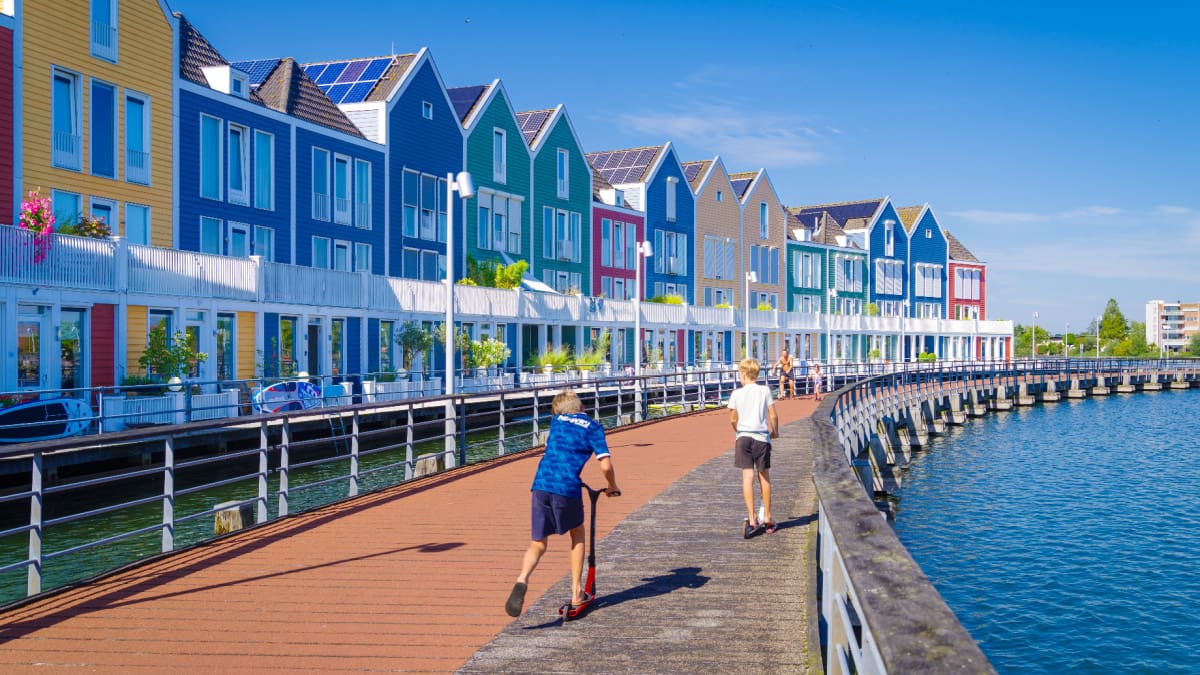 Een afbeelding van twee kinderen die spelen met een step langs het water langs een rij typisch Nederlandse huishoudens
